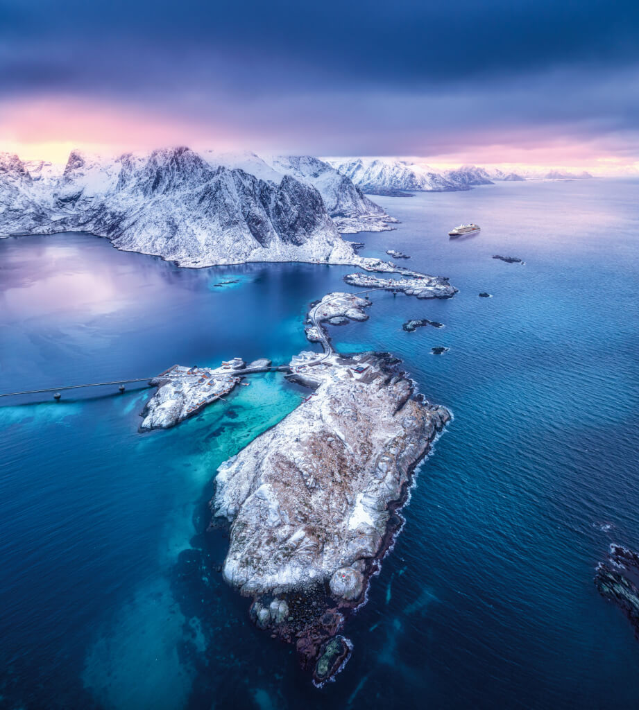 Aerial view of Hamnoy at dramatic sunset in winter in Lofoten islands, Norway. Landscape with blue sea, snowy mountains, rocks and islands, village, buildings, road, bridge, cloudy pink sky. Top view; Shutterstock ID 1869354256; purchase_order: -; job: -; client: -; other: -